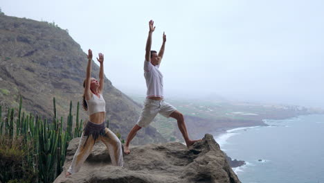 A-man-and-woman-stand-on-a-cliff,-raise-their-hands,-and-breathe-in-the-sea-air-during-yoga,-their-focus-on-the-expansive-ocean